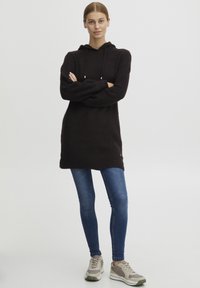Svart ribbad stickad hoodieklänning med en framficka, i kombination med blå skinnyjeans och grå sportskor. Har dragsko-detalj.