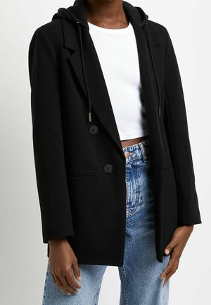 Person iført en sort blazer med hætte-detaljer, hvid cropped top og lyseblå højtaljede jeans mod en ensfarvet baggrund.