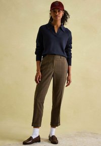 Marineblauer Pullover mit Kragen, braune karierte Hose, weiße Socken und braune Slipper. Eine kastanienrote Kappe vervollständigt das Outfit. Strukturierte Stoffdetails sichtbar.