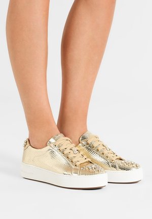 Pieds portant des sneakers dorés métalliques à tige basse avec des lacets beiges et des embellissements cloutés sur une semelle blanche, sur un fond uni.