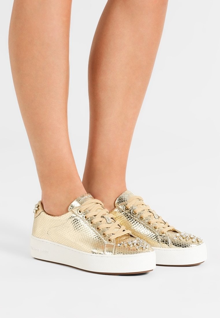 Pies que llevan zapatillas metálicas doradas de caña baja con cordones beiges y adornos de tachuelas sobre una suela blanca, con un fondo simple.
