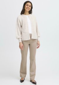 Cardigan lavorato a maglia di un beige chiaro con scollatura rotonda, maniche a palloncino e tasche frontali, abbinato a pantaloni beige e décolleté scuri a punta.