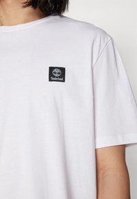 Tricou alb cu mânecă scurtă, fabricat din bumbac moale, având un eticheta Timberland de culoare neagră în formă de pătrat, cu un logo al unui copac în zona stângă a pieptului.