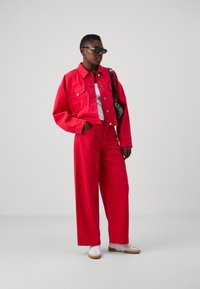 Giacca di jeans rossa con un design accorciato, abbinata a pantaloni rossi a gamba larga. T-shirt bianca sotto, accessoriata con occhiali da sole neri e una borsa piccola.