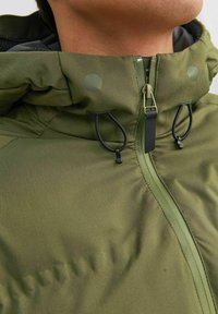 Chaqueta aislante verde con un alto cuello, que cuenta con una cremallera frontal, cordones ajustables y botones brillantes para un cierre seguro.