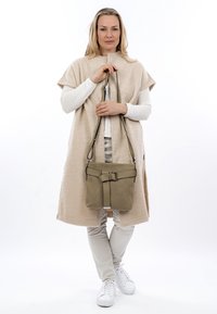 Femme portant un gilet en polaire beige, un haut blanc à manches longues, un pantalon clair, des baskets blanches, tenant un sac bandoulière vert olive avec un détail boucle.
