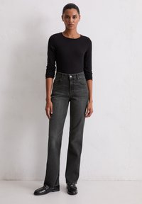 Un haut noir à manches longues associé à un jean gris foncé taille haute, avec des poches avant, une fermeture éclair et une coupe droite. Des chaussures noires complètent la tenue.