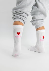 Chaussettes blanches à côtes présentant un cœur rouge et les mots "CIAO" en rose sur une chaussette et "AMOR" en rose sur l'autre.