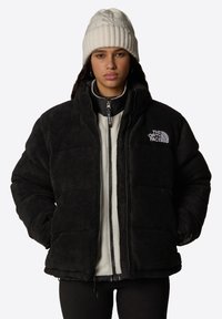 Czarna puchowa kurtka z polarowego materiału z zapięciem na zamek, stójką i detalem z logo. Nałożona na białą polarową bluzę, w połączeniu z białą wełnianą czapką.