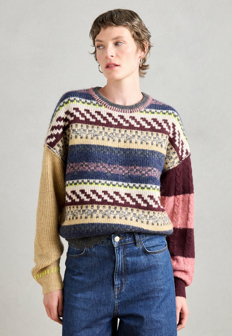 Strikkegenser med flerfargede mønstre i blått, beige og burgunder, med rund hals og varierende teksturer på ermene. Brukes sammen med blå jeans.