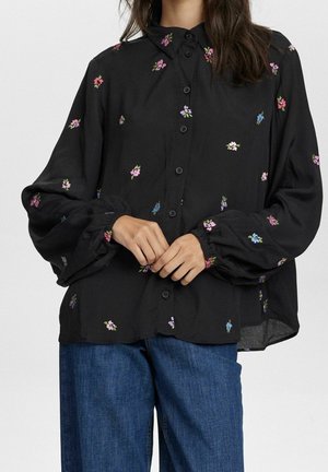 Blouse noire boutonnée avec petits motifs floraux roses, bleus et violets et manches longues bouffantes portée avec un jean bleu.