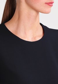 Černý top s jemnou, hladkou tkaninou, kulatým výstřihem a minimalistickým designem. Detailní záběr se zaměřuje na oblast výstřihu a ramen.