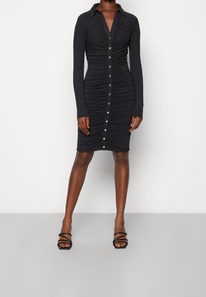 Vrouw draagt een nauwsluitende zwarte jurk met plooien, lange mouwen en knopen aan de voorkant, gecombineerd met zwarte sandalen met hak en bandjes.