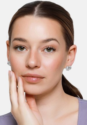 Frau mit zurückgebundenen braunen Haaren, trägt silberne Ohrringe in Knotenform, legt ihre Hand an die Wange, vor einem schlichten hellen Hintergrund.