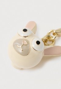 Μπρελόκ σε σχήμα κουνελιού σε κρεμ χρώμα με ροζ αυτιά, μεγάλα μάτια και χρυσές λεπτομέρειες. Κατασκευασμένο από γυαλιστερό υλικό με απαλή υφή.