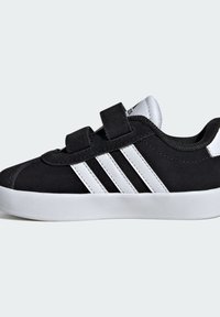 Černé semišové sportovní boty s bílými syntetickými třemi pruhy, suchými zipy a bílou gumovou podrážkou. Jsou vybaveny polstrovaným límcem pro pohodlí.