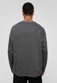 Grijze sweatshirt met lange mouwen en een gestructureerde afwerking, ronde hals, verlaagde schouders en een ruwe zoom, gedragen door een persoon van achteren.