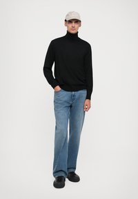 Svart polotröja, blå denimjeans och svarta sneakers. Modellen bär en ljus hatt och står mot en neutral bakgrund.