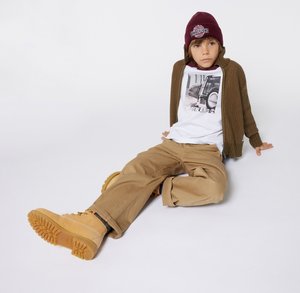 Cardigan côtelé marron, t-shirt graphique blanc, pantalon cargo beige et bottes beiges avec semelles texturées, surmonté d'un bonnet bordeaux.