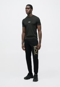 Melnas īsroku T-krekls ar "EA7" logotipu, kopā ar melnām sporta biksēm ar zelta "Emporio Armani" uzrakstu. Melni sporta apavi pabeidz izskatu.