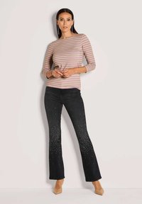 Vrouw in een gestreepte mauve en beige lange mouw top, zwarte flare jeans met sprankelende versieringen, en beige puntige schoenen, staande tegen een muur.