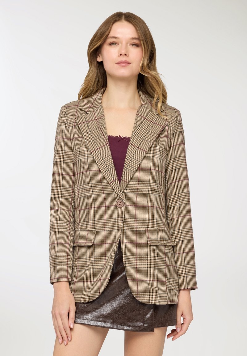 Blazer a quadri marrone con design aderente, chiusura con un solo bottone, due tasche frontali e rever a lancia. Indossato sopra una maglia color borgogna.