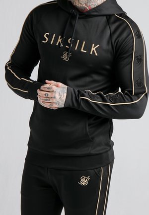 Mężczyzna w czarnej bluzie z kapturem SikSilk i pasujących spodniach z złotą lamówką i detalami logo, złożone ręce, pokazujące tatuaże na palcach i szyi.