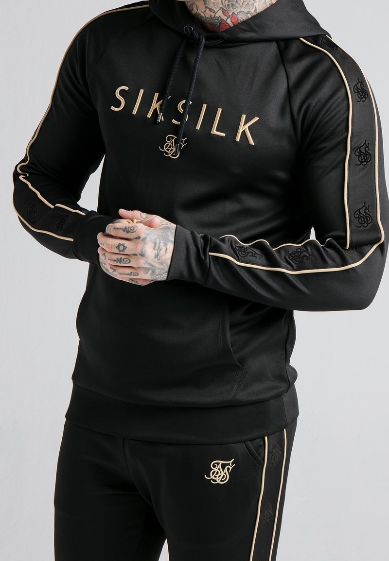 Homme portant un sweat à capuche noir SikSilk et un pantalon assorti avec des passepoils dorés et des détails de logo, mains jointes, montrant des doigts et un cou tatoués.