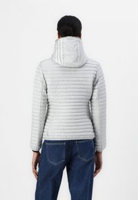 Silberne Pufferjacke mit taillierter Silhouette, horizontalen Nähten und Kapuze. Kombiniert mit blauen Jeans.