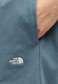 Χέρι που ακουμπά στην πλαϊνή τσέπη μπλε πλεκτού παντελονιού με το λογότυπο The North Face κεντημένο σε λευκό κοντά στην τσέπη.