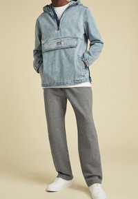 Hombre con chaqueta de denim con capucha de color azul claro, pantalones de chándal grises y zapatillas blancas, de pie contra un fondo beige liso.
