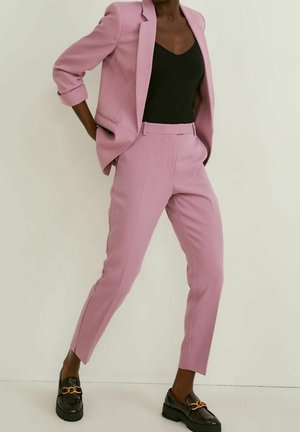 Personne portant un costume rose avec les manches retroussées, un haut noir et des mocassins noirs avec un détail de chaîne dorée, debout devant un fond clair uni.