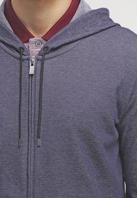 Felpa viola con zip e finitura testurizzata, dotata di una cerniera frontale, lacci regolabili e uno strato sottostante con colletto a scacchi.