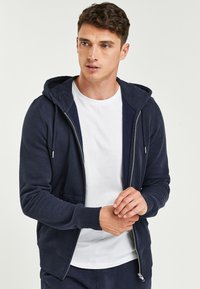 Marineblaue Zip-Hoodie aus weichem Material. Verfügt über eine Kapuze mit Kordelzug, eine frontale Tasche und gerippte Bündchen. Trägt man über einem weißen T-Shirt.