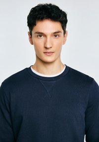 Jonge man met kort zwart haar, draagt een donkerblauwe trui met ronde hals over een wit shirt, kijkt recht vooruit tegen een effen lichte achtergrond.