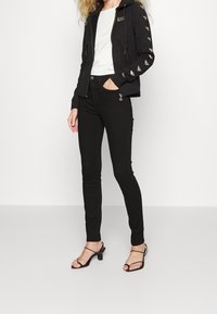 Svart zip-up hoodie med logotyp på bröstet och mönstrade ärmar, kombinerad med svarta skinny jeans och snurriga klacksandaler.