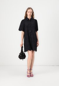 Abito camicia nero a maniche corte con motivo ad occhio, orlo scallop e chiusura con bottoni; abbinato a una borsa nera a fantasia e sandali viola con cinturini.