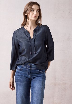 Dunkelblaue Denim-Bluse mit Dreiviertelärmeln, V-Ausschnitt und Kordelzugdetails. Kombiniert mit blauen Jeans für einen lässigen Look.