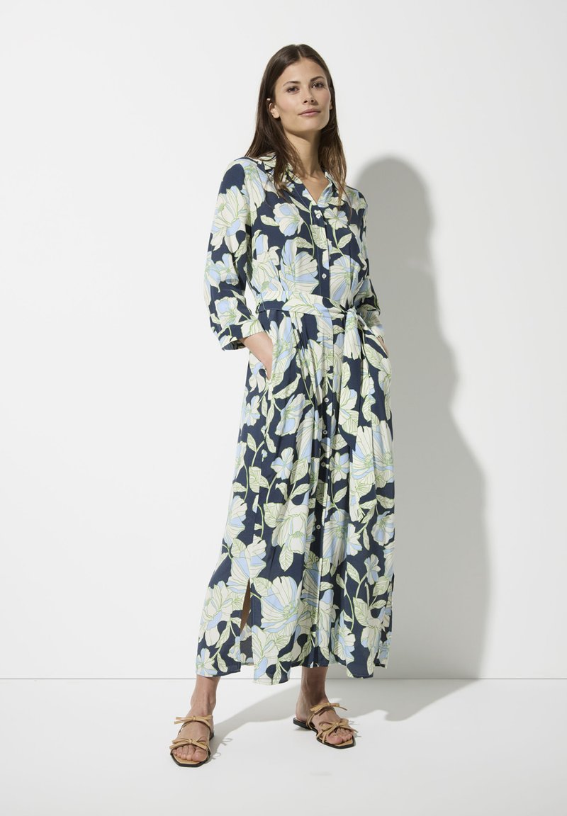 Kleid mit floralem Muster und langen Ärmeln, navy Hintergrund, große hellblaue und grüne Blätter. Verfügt über Knöpfe und eine taillierte Taille, seitliche Taschen.
