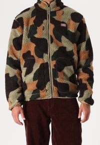 Man som bär en zipad, flerfärgad fleecejacka med camouflagemönster och stora fickor samt bruna manchesterbyxor.