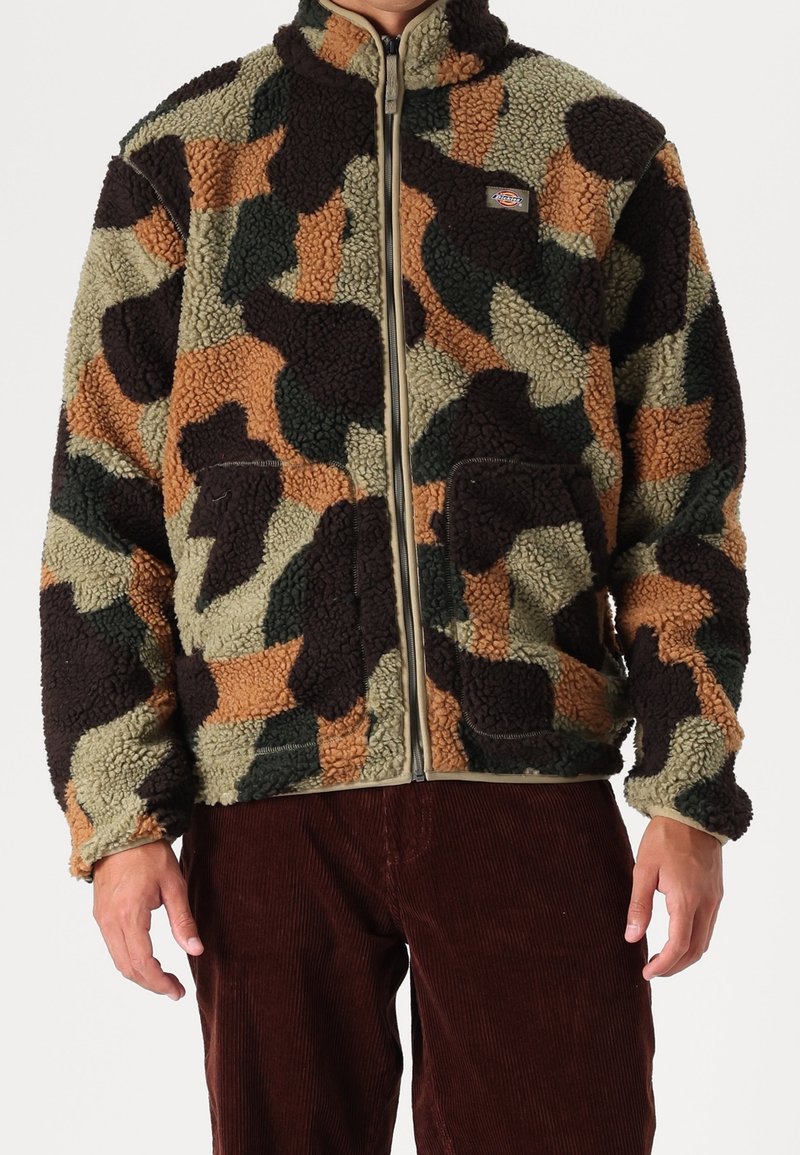 Man som bär en zipad, flerfärgad fleecejacka med camouflagemönster och stora fickor samt bruna manchesterbyxor.