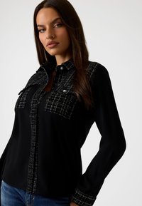 Zwarte shirt met lange mouwen en een knoopsluiting aan de voorkant. Bevat getextureerde zwart-witte ruitaccenten op de schouders en zakgebieden.