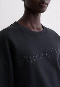Zwarte sweatshirt gemaakt van zachte stof, met een ribbelkraag en verhoogde tekst "Marc O'P" in bijpassend zwart. Close-up weergave.