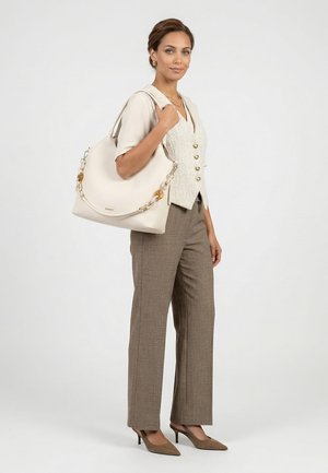 Frau trägt beige Bluse, strukturierte Weste, braune Hose und Absatzschuhe und hält eine große cremefarbene Handtasche mit Kettenriemen auf der Schulter.