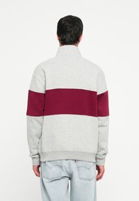 Gray sweatshirt with a maroon horizontal stripe across the back, featuring a high collar and ribbed cuffs. Soft texture, relaxed fit.