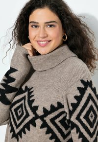 Jasnobrązowy sweter z wysokim kołnierzem, ozdobiony czarnymi geometrycznymi wzorami na rękawach. Teksturowany materiał dzianinowy o luźnym kroju.