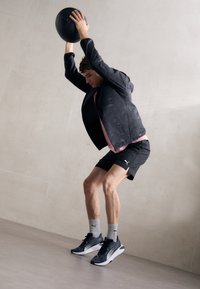 Jachetă sport neagră peste o cămașă roz, pantaloni scurți negri și șosete gri. Ține în mâini o minge de medicină neagră. Fundal gri pentru podea și pereți.