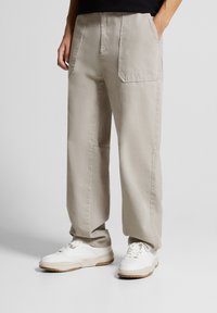 Henkilö, joka on pukeutunut löysiin beige-housuihin, joissa on suuret etutaskut, ja valkoisiin tennareihin, seisoo kädet taskuissa yksinkertaisella taustalla.