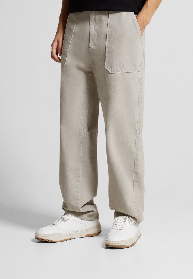 Henkilö, joka on pukeutunut löysiin beige-housuihin, joissa on suuret etutaskut, ja valkoisiin tennareihin, seisoo kädet taskuissa yksinkertaisella taustalla.