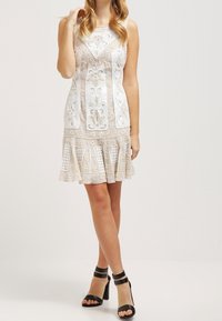 Robe blanche sans manches avec des broderies complexes, des détails en tissu transparent, un ourlet festonné et des volants superposés ; assortie à des sandales à talons hauts noires.